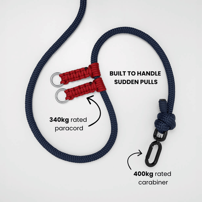 A dark navy blue rope dog lead with red paracord knots and swivel carabiner hardware, shown on a light gray background with text callouts highlighting strength ratings and load-bearing points. showing it's a dog lead thats made to handle sudden pulls