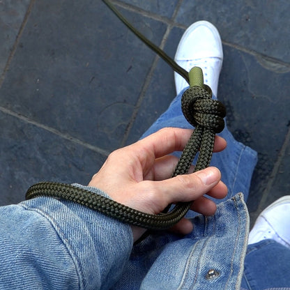 Hand holding a green swivel slip lead for dogs with a knot, wearing blue jeans and white sneakers on a dark tiled floor.